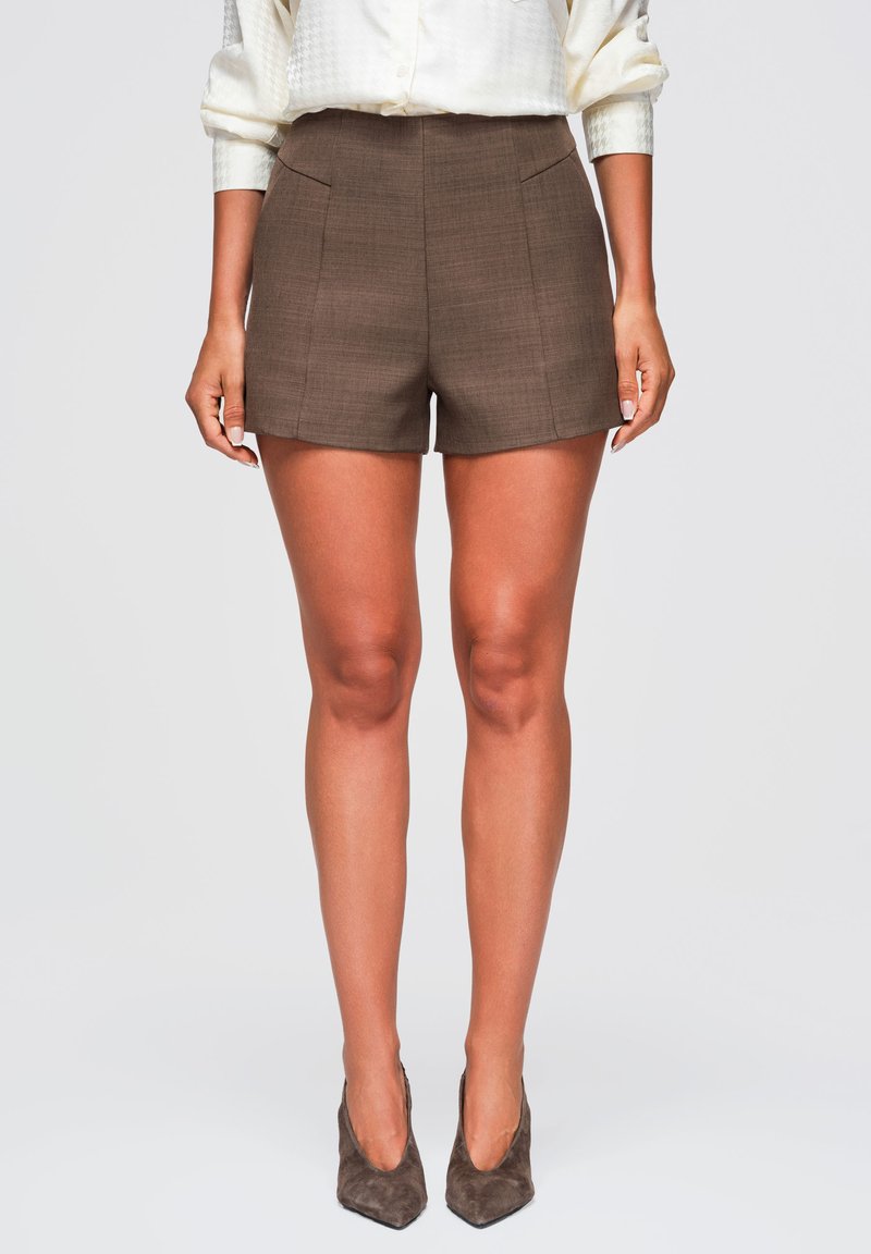 Femme portant un short marron cintré, une blouse blanche boutonnée avec des manches retroussées, et des escarpins marron en daim à bout pointu, debout, les mains détendues.