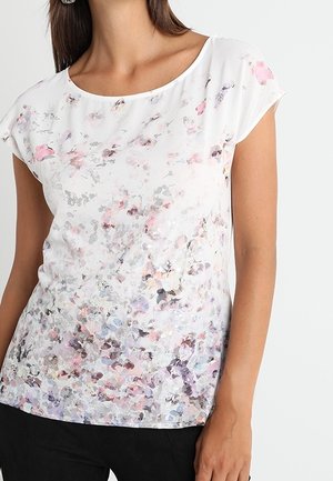 Mujer que lleva una blusa blanca de manga corta con un sutil estampado floral abstracto en rosa, gris y púrpura, combinada con pantalones negros.