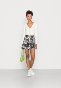 Top cropped branco com detalhes em ribana e frente com atacadores, saia mini com padrão de zebra, bolsa verde e ténis brancos. Fundo plano para contraste.