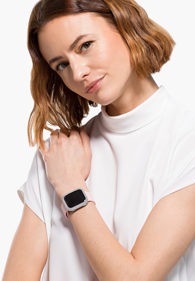 Montre intelligente avec un écran tactile carré noir, une lunette argentée ornée de strass et un bracelet en silicone rose pâle, portée au poignet.