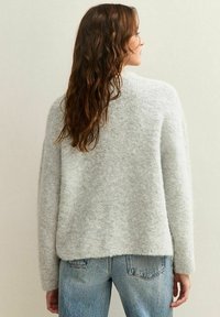 Vrouw met lang bruin haar in een lichte grijze fluffy trui en blauwe jeans, met haar rug naar de camera gekeerd tegen een effen lichte achtergrond.
