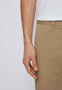 Béžové bavlněné kalhoty s plochým předním dílem, které mají boční kapsy a hladkou texturu. Ruka spočívá na boku kalhot.