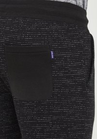 Fekete és szürke mintázatú joggers, bordás derékpánttal, oldalsó zsebbel és egy kis lila címkével. Texturált anyag vízszintes csíkokkal.