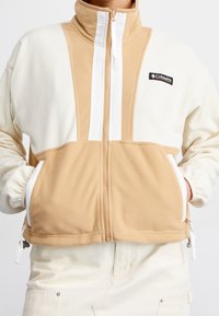 Fleecejacka med hög krage, med paneler i beige och vitt, dragkedjestängning, två framfickor och en logotyp på vänster sida.