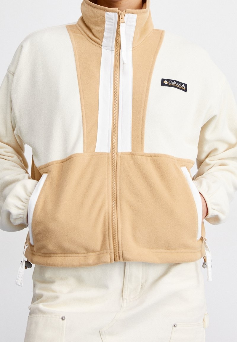Fleecejacka med hög krage, med paneler i beige och vitt, dragkedjestängning, två framfickor och en logotyp på vänster sida.