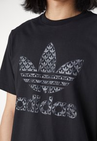 Černé bavlněné tričko s velkým šedým logem trefoil s vzorovaným designem a nápisem „adidas“ ve stejném odstínu šedi ve spodní části.