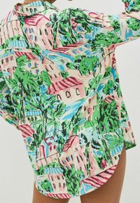 Chemise colorée avec un imprimé de paysage urbain abstrait, mettant en avant des maisons avec des toits rouges, des arbres verts et un ciel bleu, portée par une personne avec le bras levé.