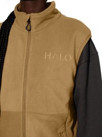 Close-up af person iført tan fleecevest med "HALO" broderi over sort langærmet skjorte, med frontlynlås og sidelomme.