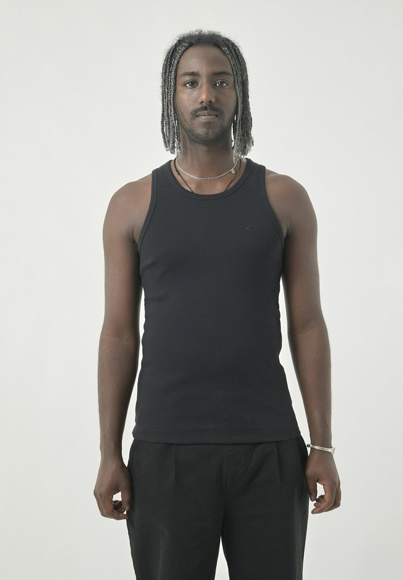 Schwarzes geripptes Tanktop mit rundem Ausschnitt, figurbetontem Design und dünnen Trägern, getragen mit schwarzen, locker sitzenden Hosen.