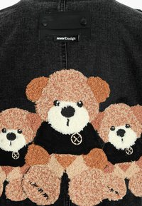 Chaqueta de mezclilla negra con aplicaciones de ositos de peluche marrón. Los osos llevan camisetas negras, con detalles de textura y bordado notables.