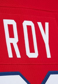 Tessuto rosso testurizzato con lettere bianche cucite "ROY" e una vista parziale di un numero bianco e blu sottostante.