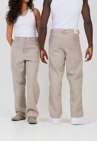 Pantaloni bej cu croială largă, dintr-un material neted, având buzunare laterale și petice la spate, purtați cu încălțăminte în nuanțe neutre.