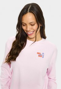 Cămașă roz cu mânecă lungă, guler rotund, având text multicolor „mare” și „soare” și un grafic cu un cal de mare pe partea stângă a pieptului.