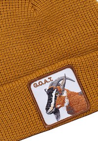 Cappello di lana color senape con un patch che presenta un design di una capra ricamato e il testo "G.O.A.T." in grassetto sopra l'illustrazione.