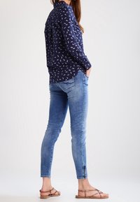Camisa de botones azul marino con un patrón floral blanco, combinada con jeans de mezclilla azul claro con cremalleras en los tobillos y sandalias marrones.