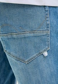Lichtblauwe denim jeans hebben een gestikte achterzak met een gebogen ontwerp en een klein wit label met blauwe stippen aan de zijkant.