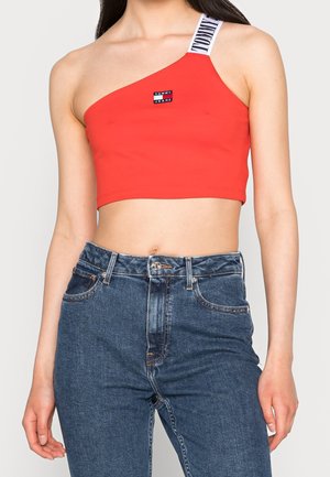 Kobieta w czerwonym krótkim topie na jedno ramię z białym paskiem Tommy Jeans oraz wysokim stanem niebieskich dżinsów na jednolitym tle.