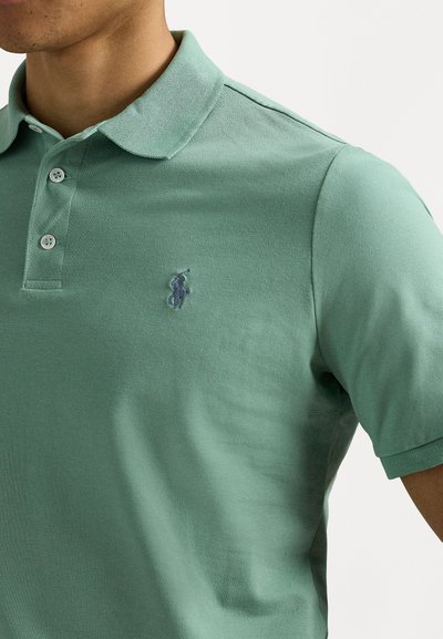 Zelena polo majica od teksturiranog pamuka. Ima klasičan ovratnik, tri dugmeta i mali plavi izvezeni logo na lijevoj strani prsâ.