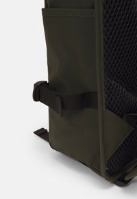 Olivenfarget ryggsekk laget av holdbart materiale med en teksturert mesh-panel, justerbar stropp og en sidelomme for ekstra oppbevaring.