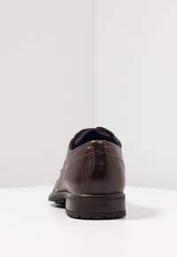 Chaussure habillée en cuir marron foncé avec des accents de couture rouge, vue de dos sur une surface blanche et un fond clair.