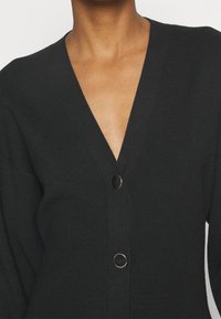 Cardigan noir à côtes avec un col en V et trois boutons sur le devant, présentant une texture douce et des manches légèrement surdimensionnées.