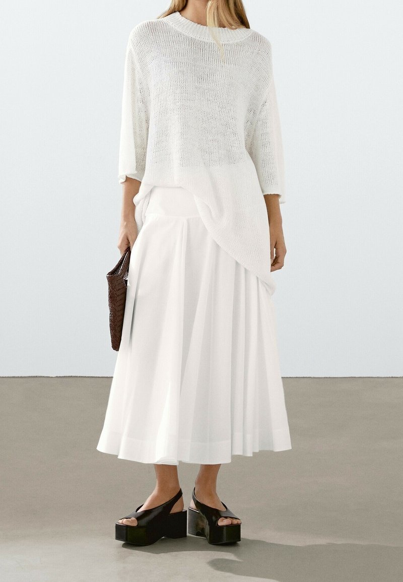 Witte brei top met een losse pasvorm en driekwart mouwen, gecombineerd met een lange, vloeiende witte rok. Zwarte plateau-sandalen en een bruine gevlochten tas.