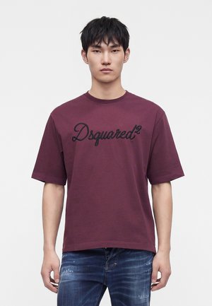 Junger Mann, der ein einfarbiges dunkelrotes T-Shirt mit kurzen Ärmeln mit dem Text "Dsquared2" und blaue Jeans trägt, vor weißem Hintergrund stehend.