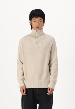 Beige gestreifter Pullover mit hohem Kragen und halben Reißverschluss, ausgestattet mit einem strukturierten Strickmuster und taillierten Bündchen, kombiniert mit schwarzen Hosen.