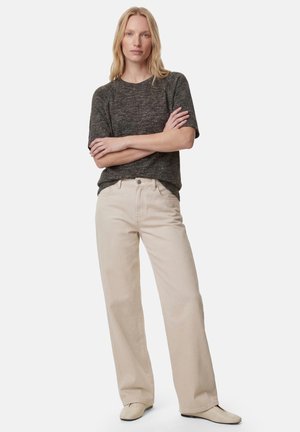 Donna bionda in piedi con le braccia incrociate, indossa una maglietta grigio scuro a maniche corte, pantaloni beige a gamba larga e scarpe beige chiaro su sfondo uniforme.