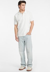 Polo shirt bianco a maniche corte, jeans azzurri chiari e sneakers beige con dettagli bianchi, in piedi su uno sfondo neutro.