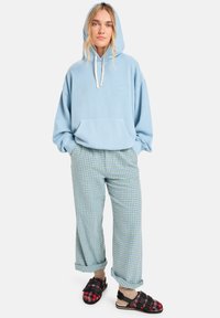 Ljusblå huvtröja med ficka framtill, kombinerad med grön och blå rutiga byxor samt svarta sandaler med röda detaljer och spännen.