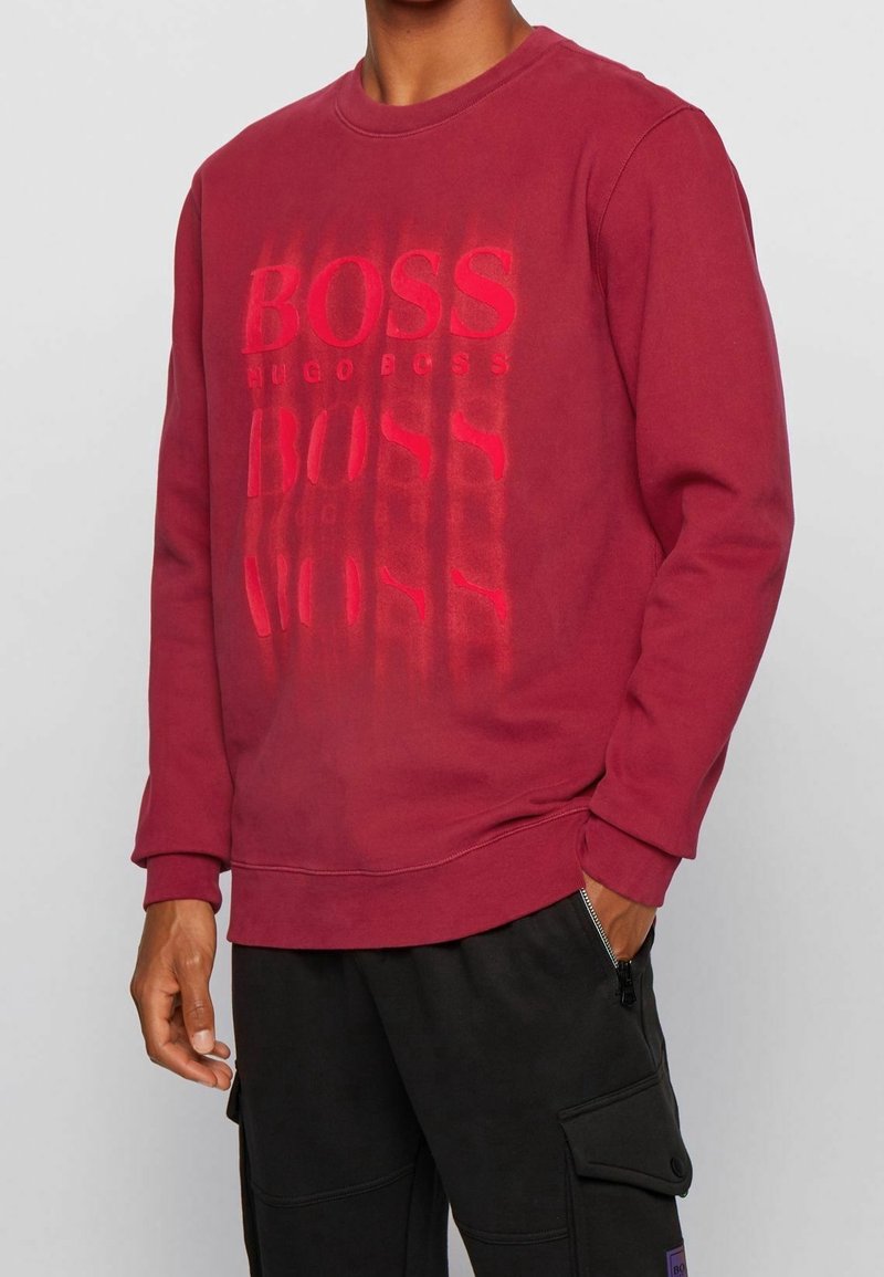 Felpa bordeaux con scollo rotondo, maniche lunghe e una stampa testuale "BOSS" in diverse tonalità. Materiale morbido con una vestibilità rilassata.
