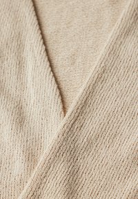 Beige texturerad tyg med ribbat mönster, som har en djup V-ringning och överlappande paneler. Mjuk och stickad material.