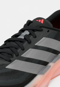 Μαύρο και γκρι αθλητικό παπούτσι Adidas με τρεις αντανακλαστικές ρίγες, μαύρα κορδόνια και κόκκινο λογότυπο στη γλώσσα, σε λευκό φόντο.