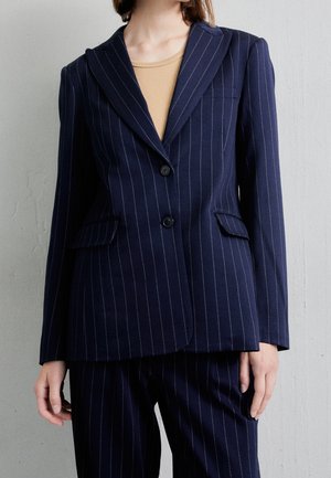 Frau trägt ein dunkelblaues Nadelstreifen-Blazer mit zwei Knöpfen über einem hellbraunen Top, kombiniert mit passenden Nadelstreifen-Hosen.