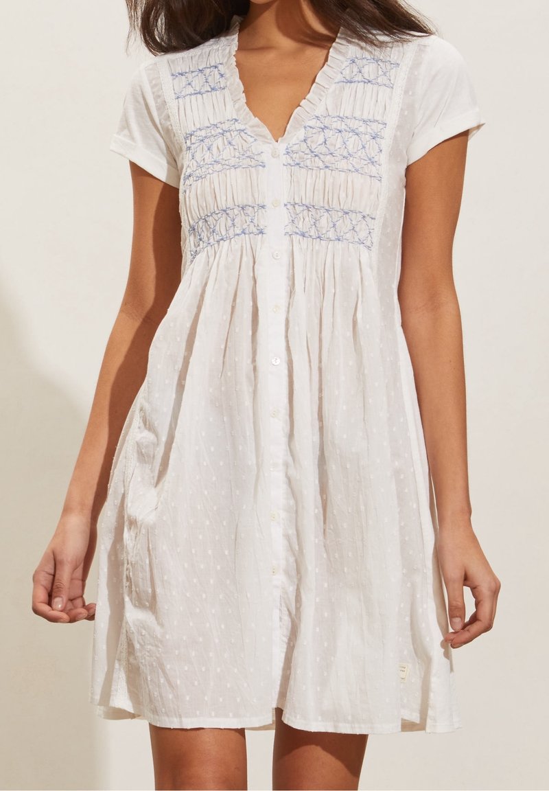Abito bianco a maniche corte con motivi ricamati blu sul petto, chiusura con bottoni sul davanti e una sottile texture a pois, indossato da una persona in piedi.