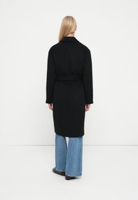 Cappotto nero con cintura, colletto, maniche lunghe e silhouette dritta, abbinato a jeans a gamba larga blu chiaro.