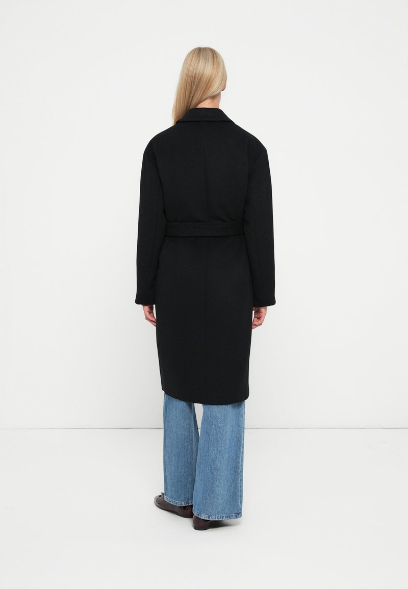 Svart bältejacka med krage, långa ärmar och rak siluett, i kombination med ljusblå vida jeans.