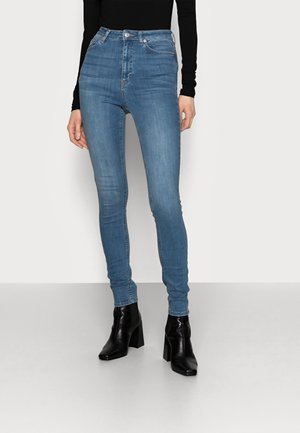 Schmal geschnittene blaue Jeans aus Denim, kombiniert mit einem schwarzen Langarmshirt und schwarzen Stiefeletten mit Absatz, stehend vor einem schlichten, hellen Hintergrund.