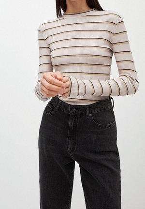 Femme portant un haut à manches longues beige et noir à rayures, rentré dans un jean noir taille haute, mains jointes au niveau de la taille.