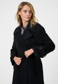 Svart trench coat i mjukt tyg, med dubbelknäppt design, stora slag, knappar och ett avtagbart bälte i midjan.