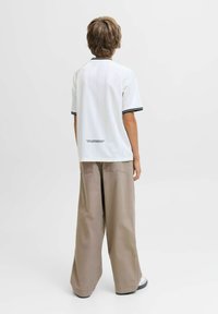 Persona con capelli corti sta in piedi di spalle, indossando una maglietta bianca a maniche corte con bordi neri e pantaloni beige larghi con tasche.