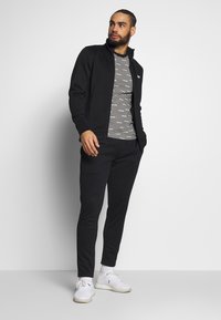 Črna zip-up jakna preko črno-belih črtastih majic, v kombinaciji s črnimi trenirko hlačami in belimi športnimi čevlji. Gladek material in prilegajoča zasnova.