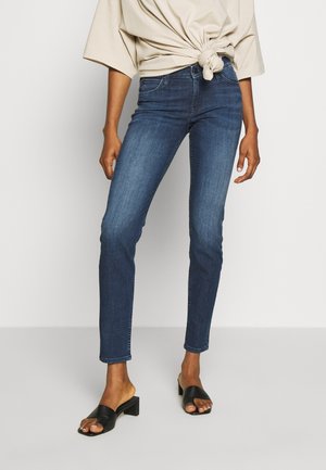 Persoon die donkerblauwe skinny jeans draagt, een beige vastgeknoopte blouse en zwarte sandalen met blokhak en open tenen, staand tegen een effen achtergrond.