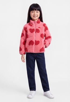 Jeune fille souriante portant une veste polaire rose avec des cœurs rouges, un jean bleu foncé et des baskets blanches, debout devant un fond blanc.