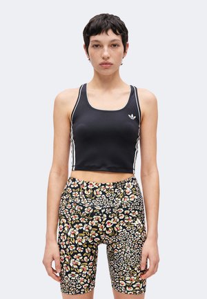 Junge Person mit kurzen dunklen Haaren, die ein schwarzes, kurzes Tanktop und hoch taillierte Fahrradhosen mit Blumenmuster vor schlichtem Hintergrund trägt.