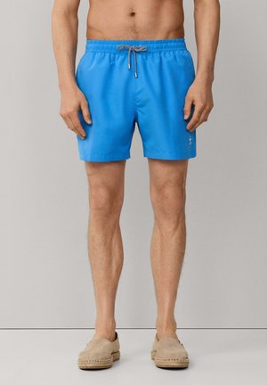 Homme portant un short de bain bleu vif avec un cordon de serrage et des espadrilles beiges, debout devant un fond uni.