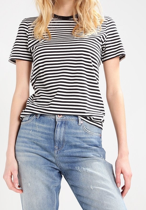 Femme portant un t-shirt à manches courtes rayé noir et blanc, rentré dans un jean bleu clair déchiré, debout devant un fond uni.