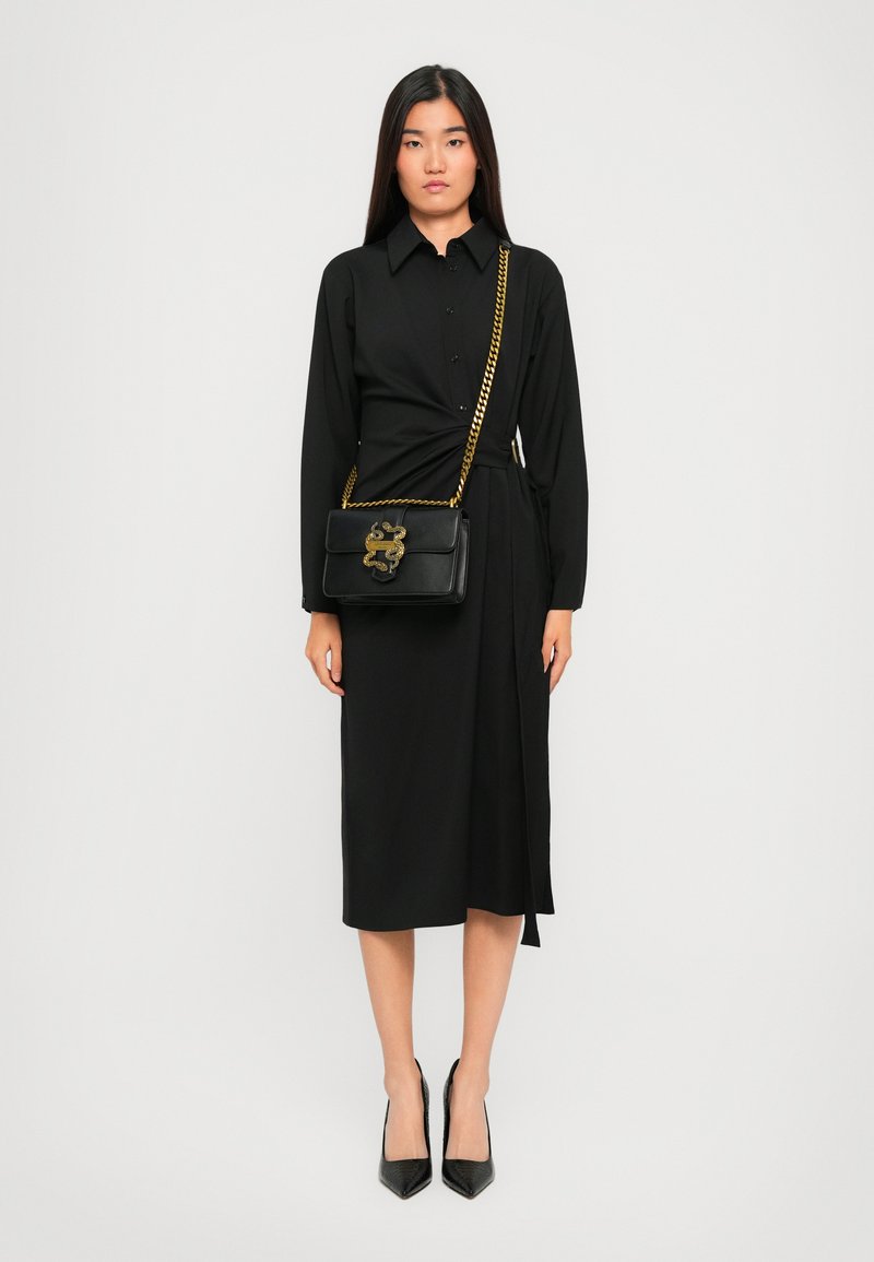 Femme en robe chemise midi à manches longues noires portant des escarpins noirs à bout pointu et un sac à épaule noir avec une chaîne dorée et un détail serpent.