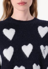 Maglione lavorato a maglia blu navy con motivi a cuore bianco peloso; presenta un colletto rotondo e un tessuto morbido e texturizzato.